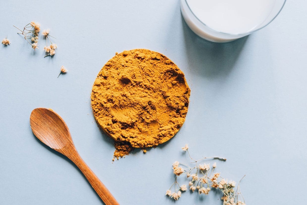 Overhead shot of turmeric powder and a wooden spoon on a light blue background.