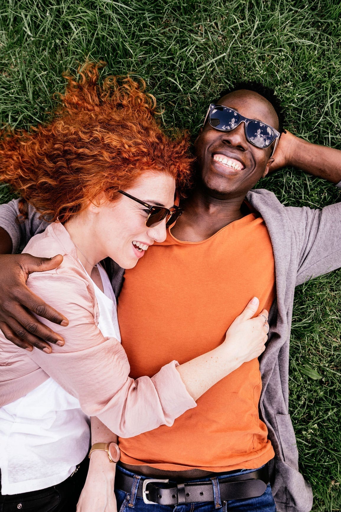 From above shot of laughing black man embracing a redheaded woman while cuddling on green grass in summer.