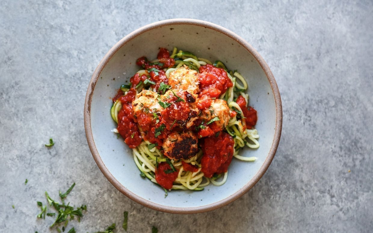 An overhead shot of a bowl filled with zucchini noodles, marinara sauce and meatballs on a grey countertop.