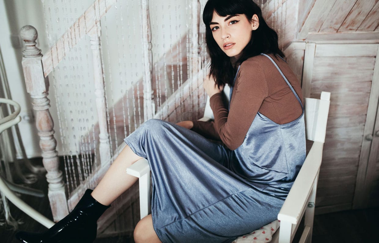 A young woman wearing a light blue silk dress with a brown long sleeve shirt sits on a white wooden chair.