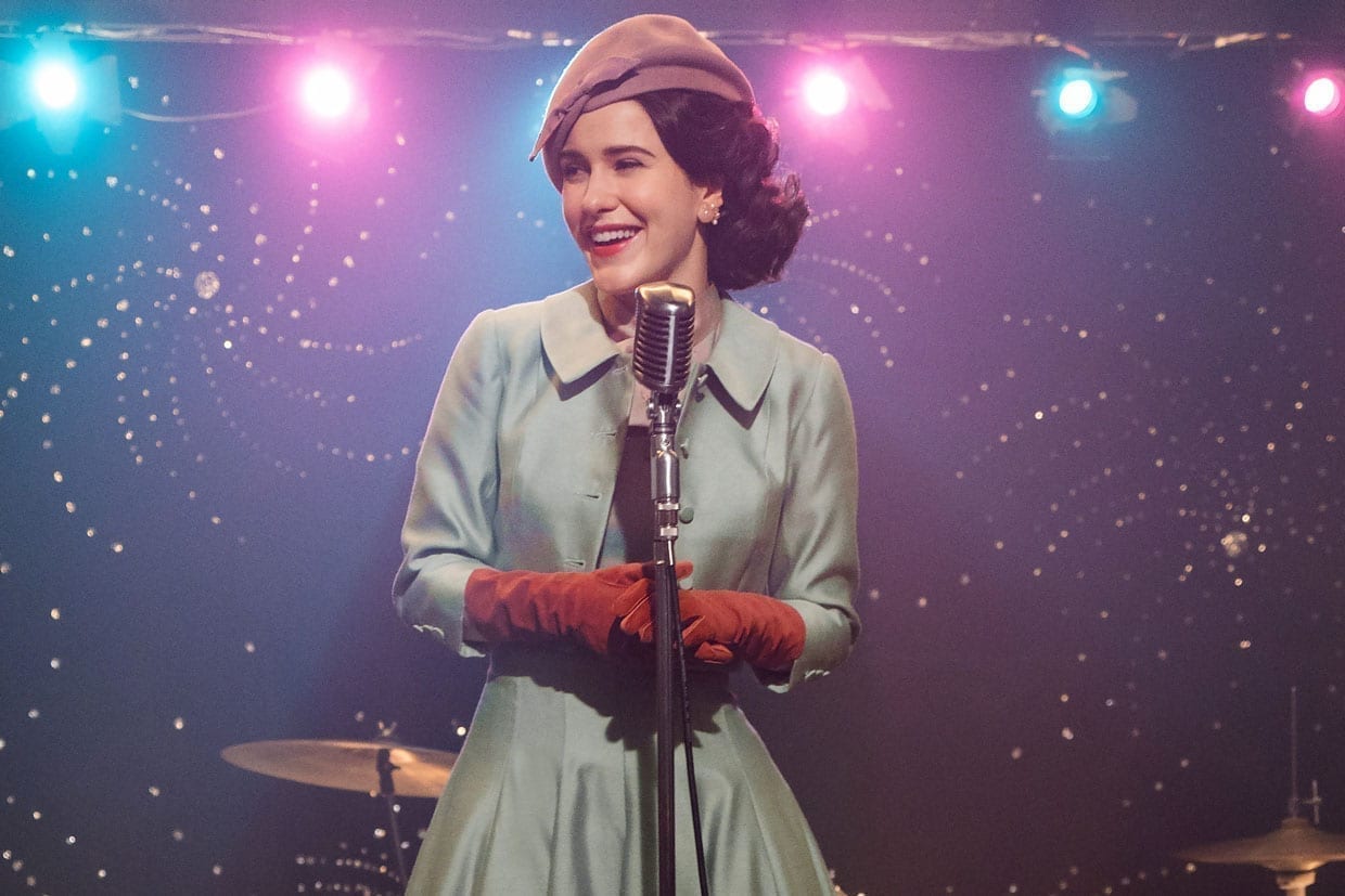 Rachel Brosnahan wears a light blue coat, red gloves, a blush hat and pink lipstick in a scene from The Marvelous Mrs. Maisel.