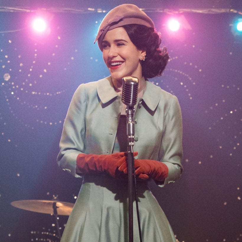 Rachel Brosnahan wears a light blue coat, red gloves, a blush hat and pink lipstick in a scene from The Marvelous Mrs. Maisel.
