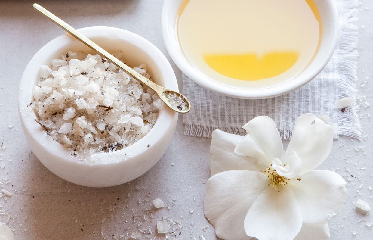 Close up of bath salts in a ceramic dish, oil in a ceramic dish and a white flower on a counter.