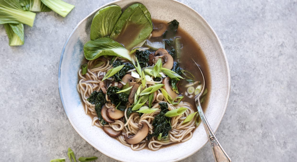 Miso noodle soup with mushrooms and kale in a white bowl with a silver spoon on a grey kitchen counter.