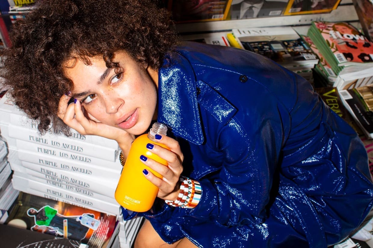 A woman with curly hair holding a yellow bottle of Dirty Lemon.