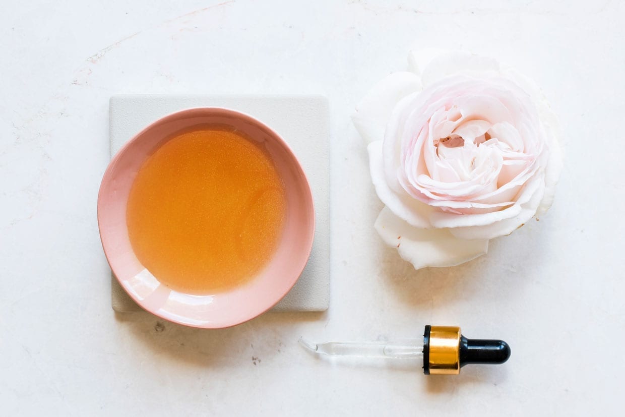 A homemade oil based hair mask sits in a bowl on a counter next to a pink rose and a dropper.
