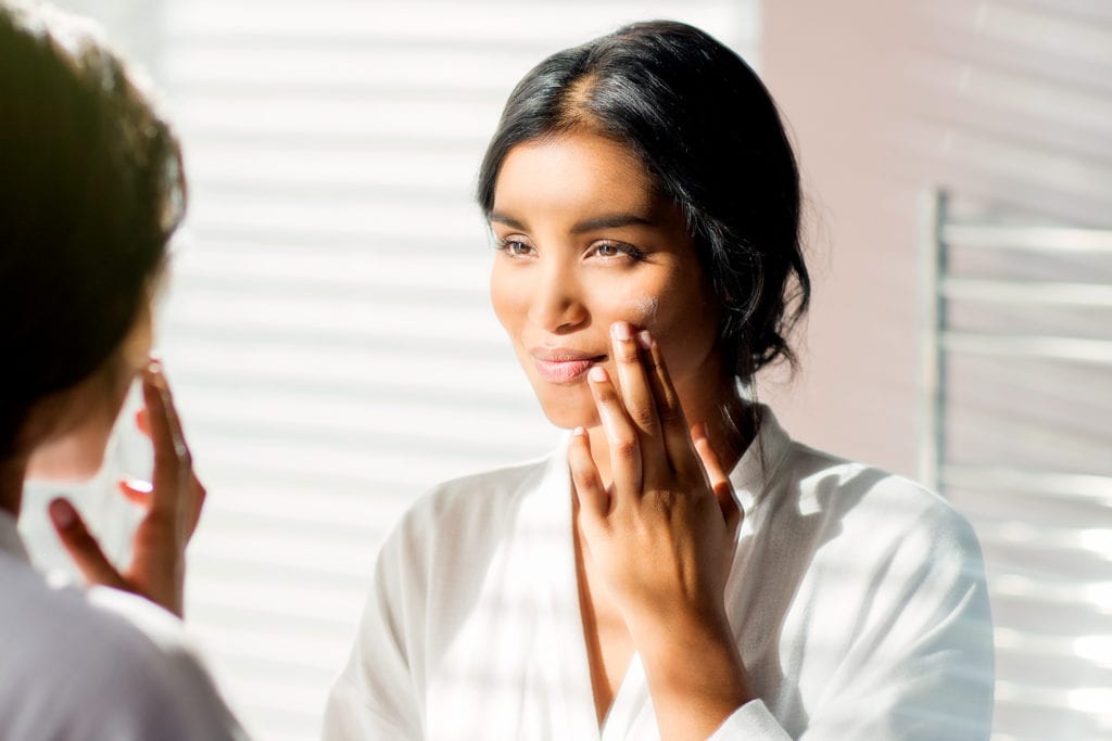 A woman applying skincare products in the bathroom mirror.