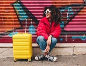 A woman in a red down jacket and jeans sits on a curb with her yellow suitcase.