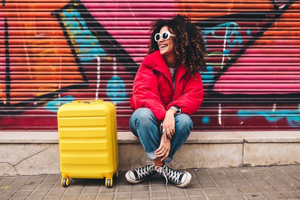 A woman in a red down jacket and jeans sits on a curb with her yellow suitcase.
