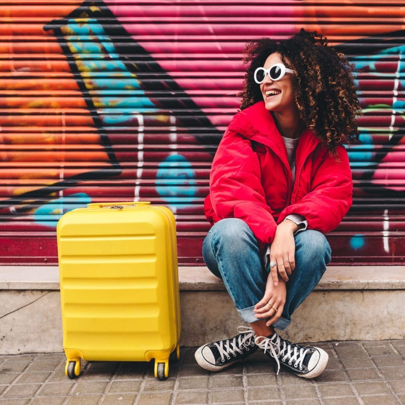 A woman in a red down jacket and jeans sits on a curb with her yellow suitcase.
