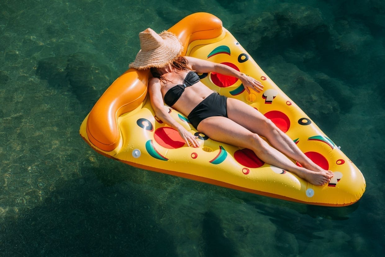 A woman wearing a natural straw hat and black bikini floats on an inflatable pizza at the beach.