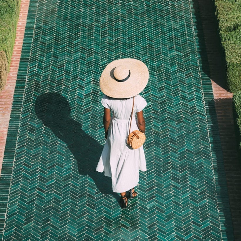 A black woman in a white dress and sunhat walking on a green tiled sidewalk.