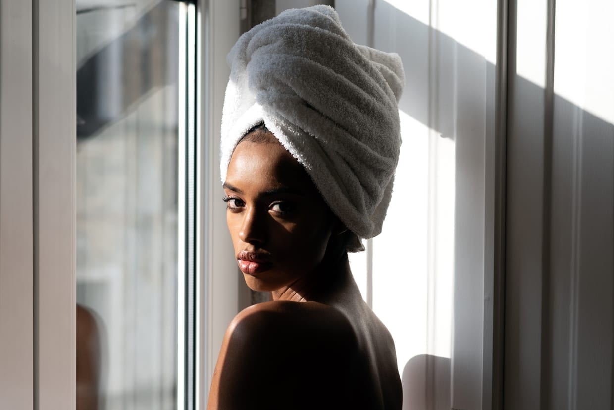 A black woman wrapped in a towel standing by a window looking at the camera.