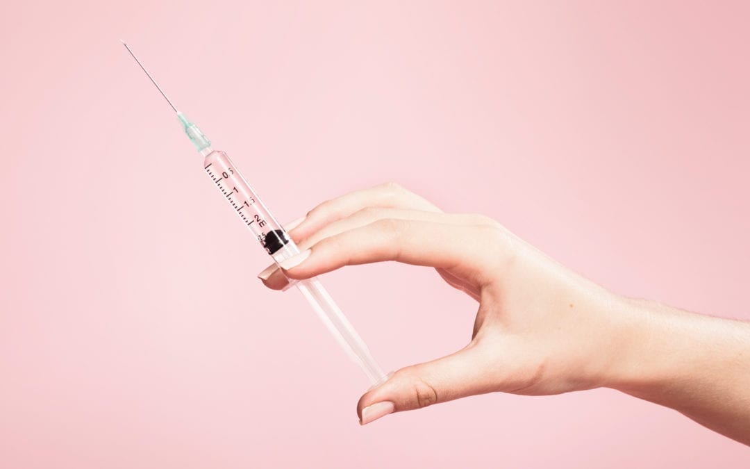 A hand holding a syringe in front of a pink background.