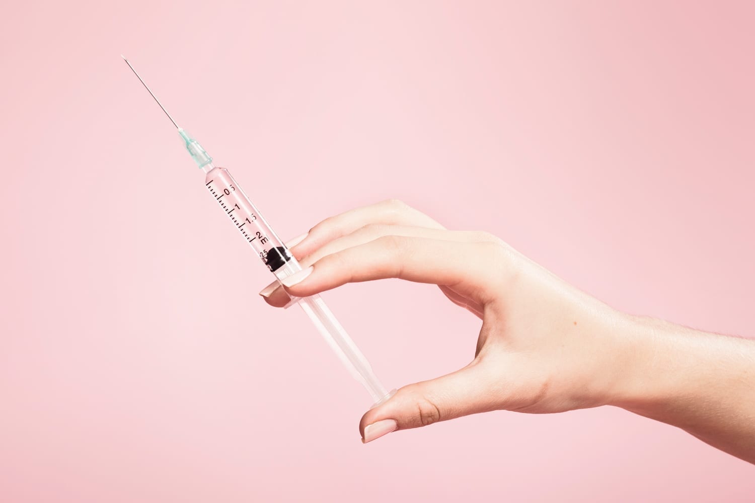 A hand holding a syringe in front of a pink background.