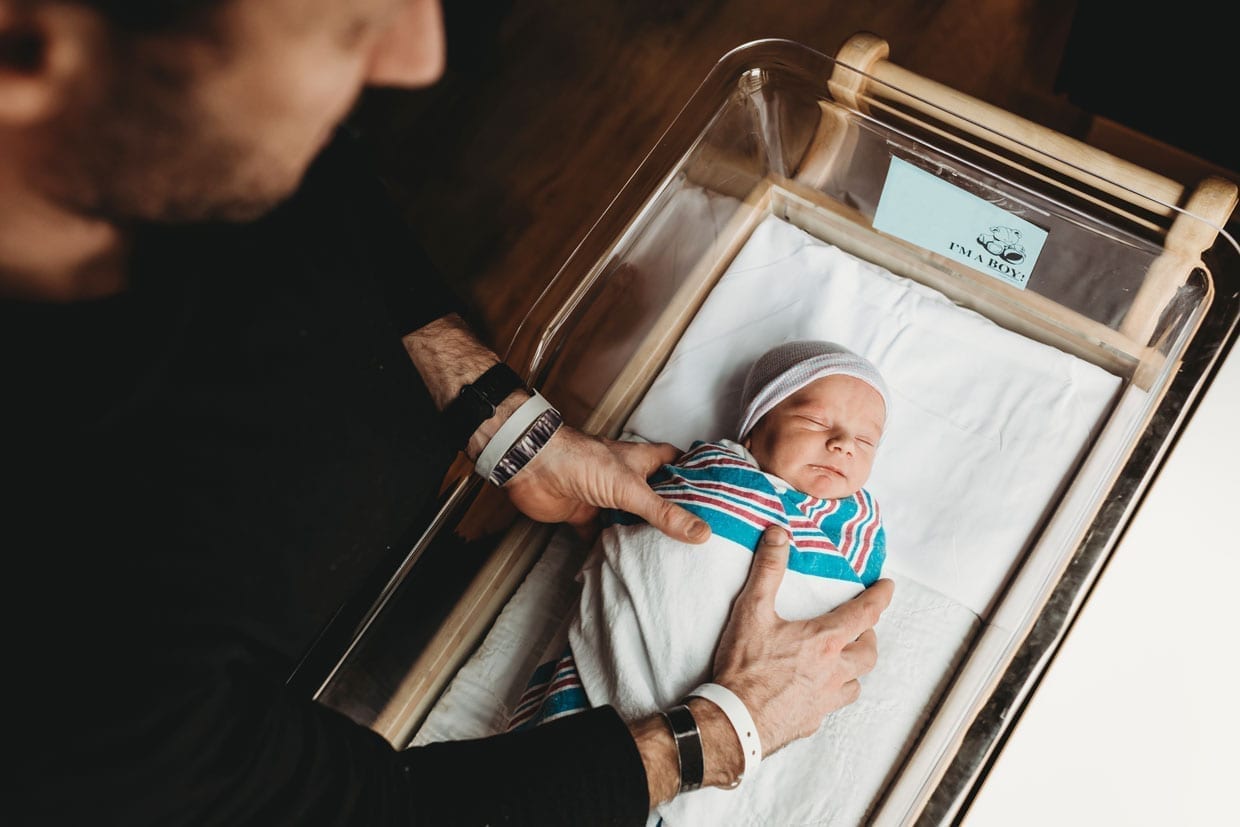 A father's hands reaching in to hospital bassinet to pick up newborn swaddled baby.