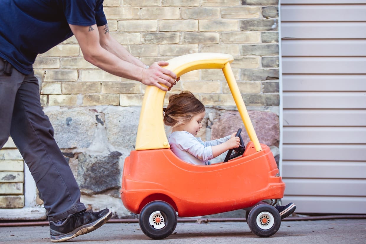 A dad pushing his daughter's toy car while she drives.