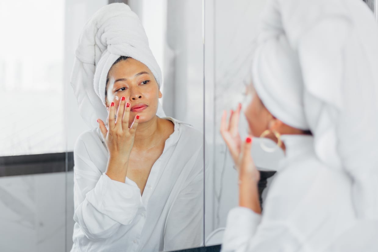 A woman applying cream on her face in front of a mirror.