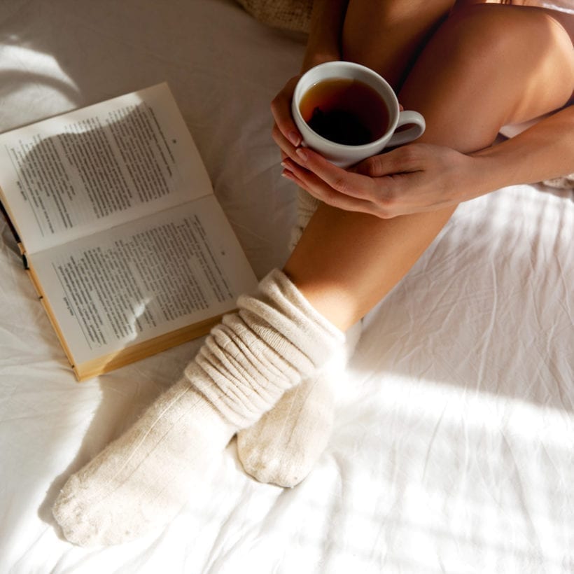 A woman wearing knitted socks holds a cup of tea in bed.