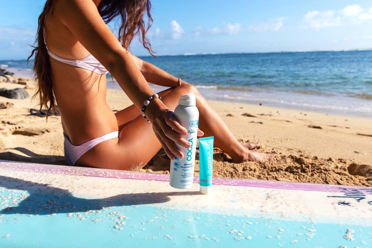 A woman next to a surfboard reaches for a spray bottle of SPF.