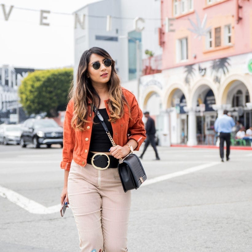 Nikki Sachdeva wearing orange jacket, Chanel bag, striped pants, sunglasses in front of the Venice Beach sign, April 22, 2018 in Los Angeles.