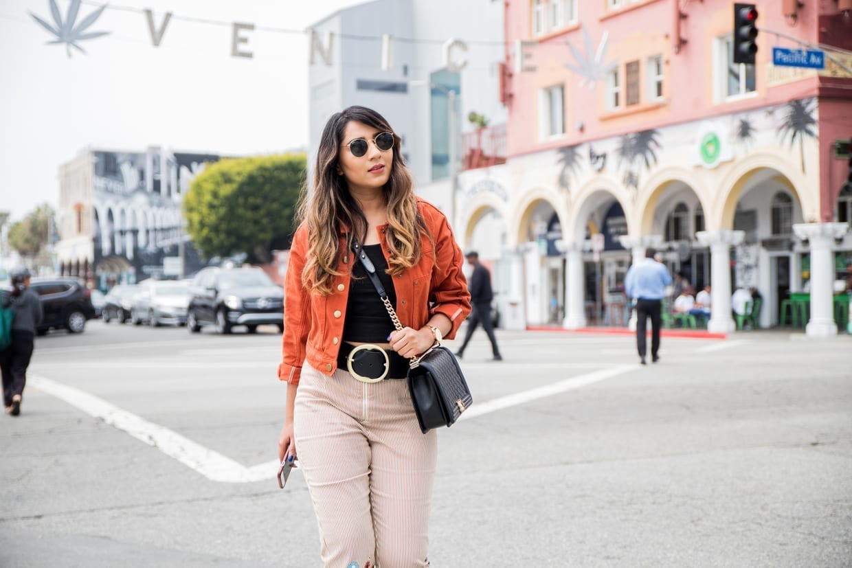 Nikki Sachdeva wearing orange jacket, Chanel bag, striped pants, sunglasses in front of the Venice Beach sign, April 22, 2018 in Los Angeles.