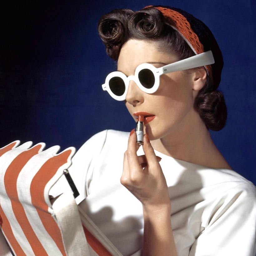 Model Muriel Maxwell in white sunglasses putting on lipstick, wearing red-white-and-blue turban, andv holding a red-and-white striped bag.