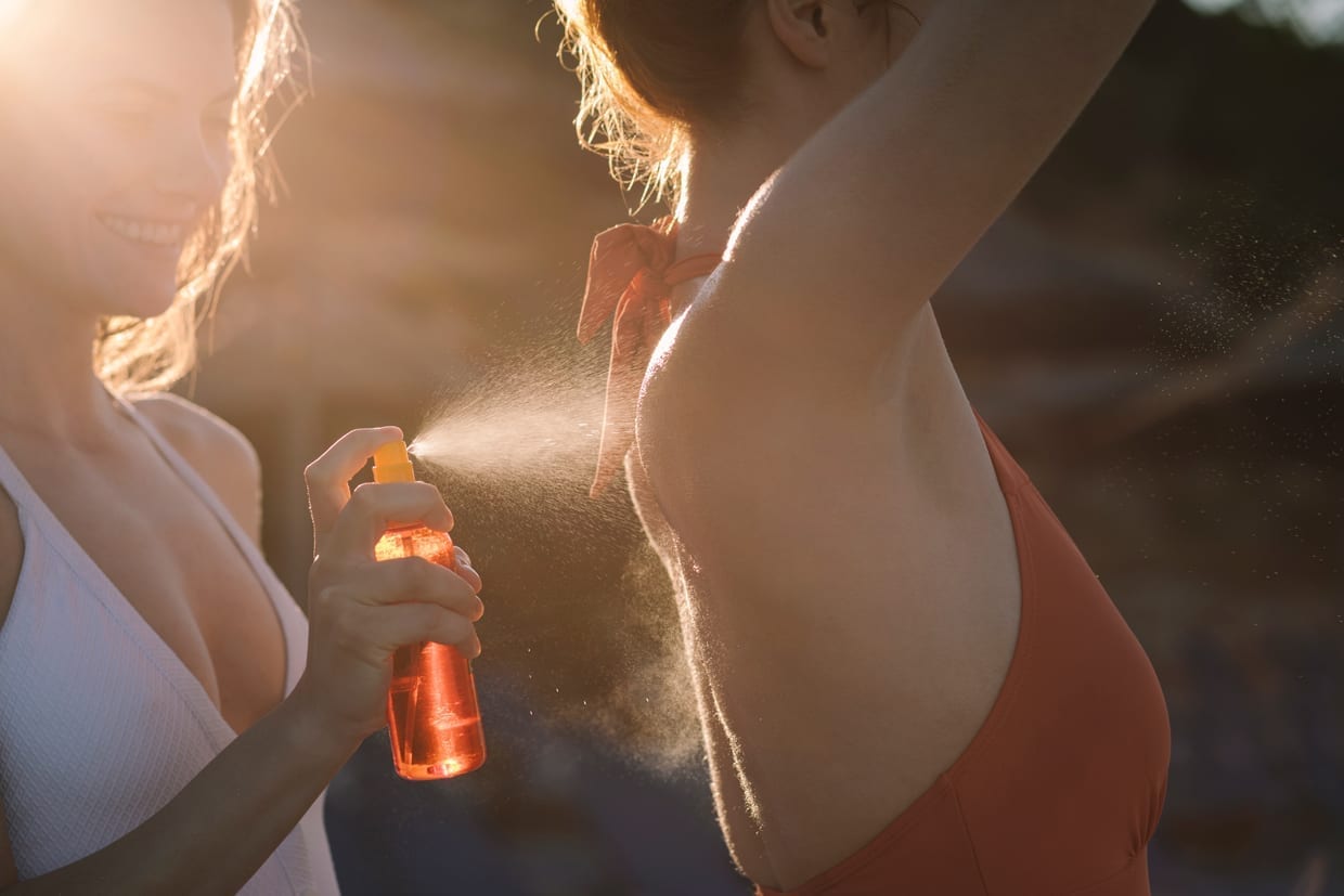 A woman sprays sunscreen on another woman's back.