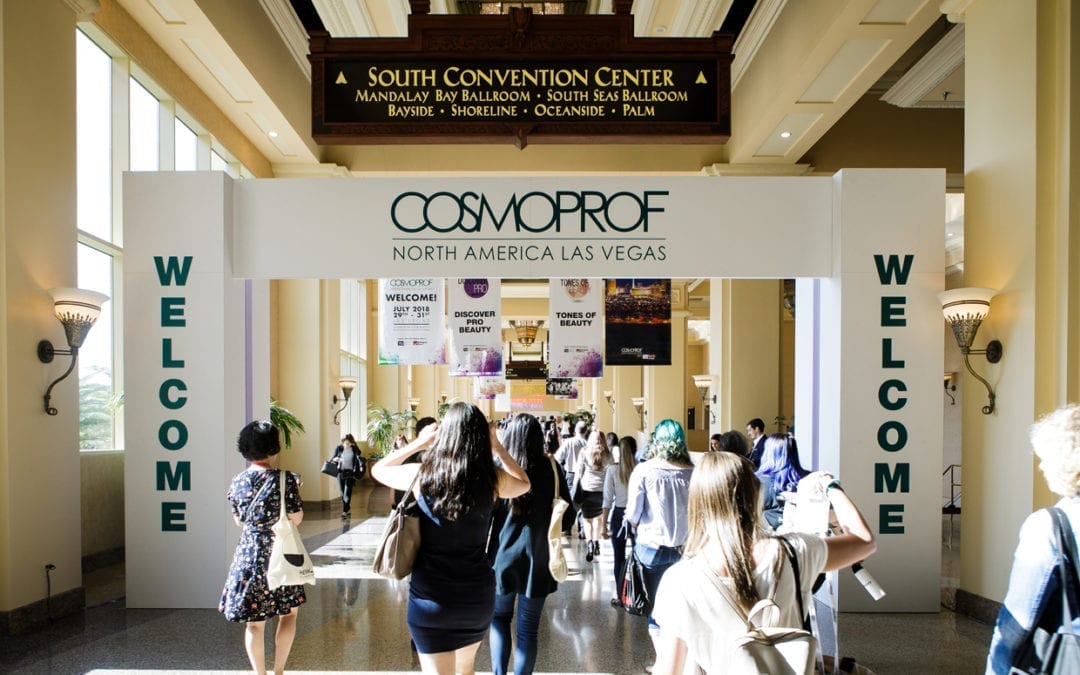 Several people walk under a white arch that reads "Welcome" on both sides and in the middle "CosmoProf North America Las Vegas."