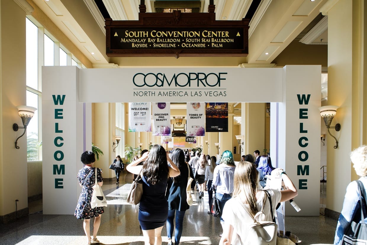 Several people walk under a white arch that reads "Welcome" on both sides and in the middle "CosmoProf North America Las Vegas."