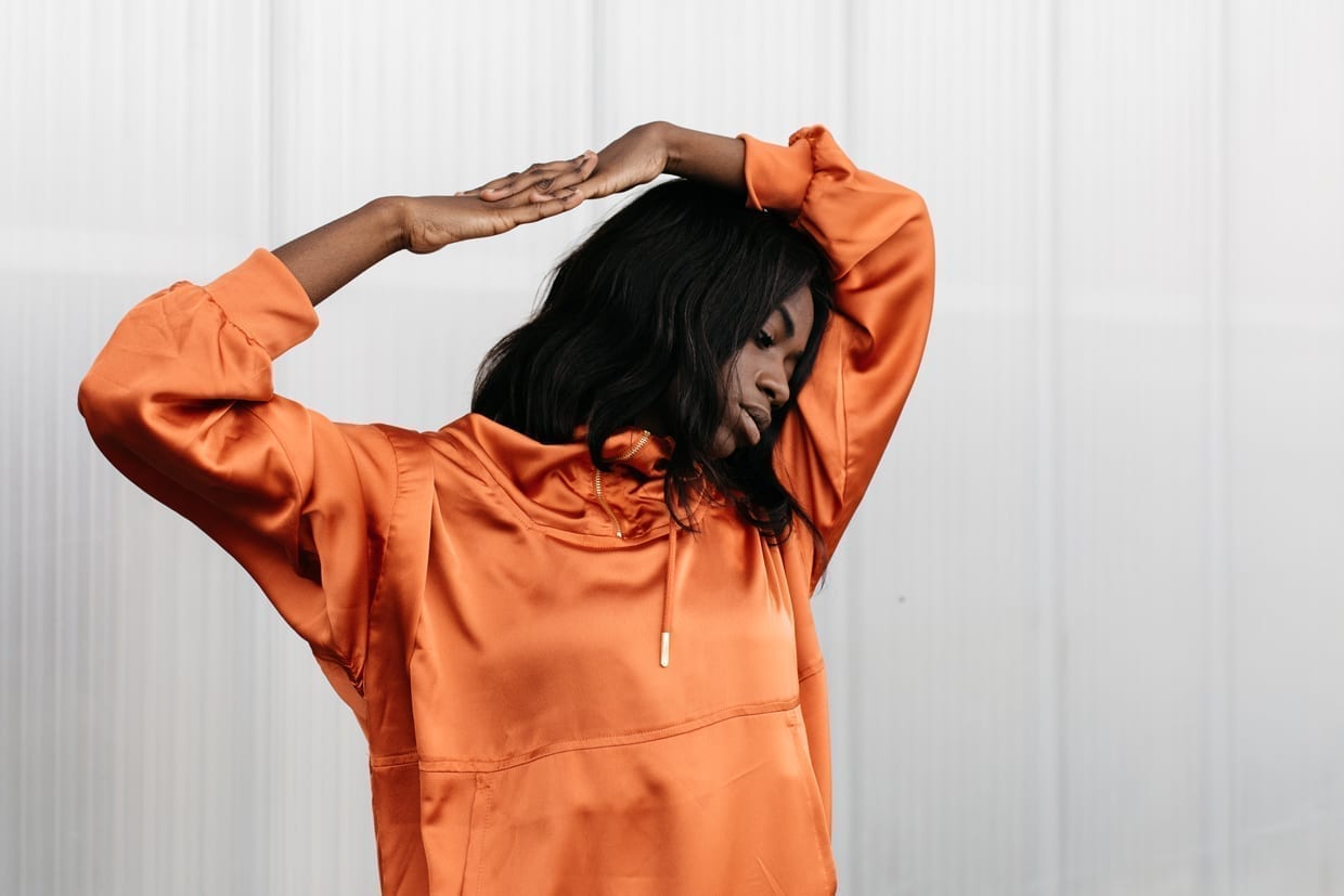 A pensive black woman in an orange hoodie holding her hands above her head and looking down.