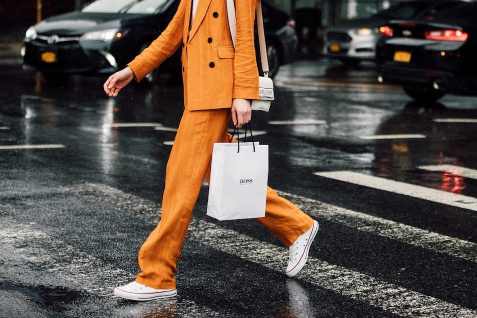 A woman in an orange suit walks across a crosswalk holding a white shopping bag.