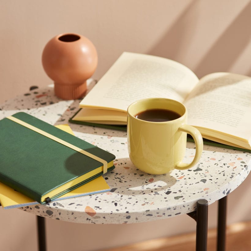 Close-up of pale yellow mug of hot coffee, green planner and an open book on round marble table in sunlight.