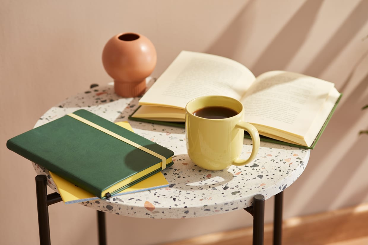 Close-up of pale yellow mug of hot coffee, green planner and an open book on round marble table in sunlight.