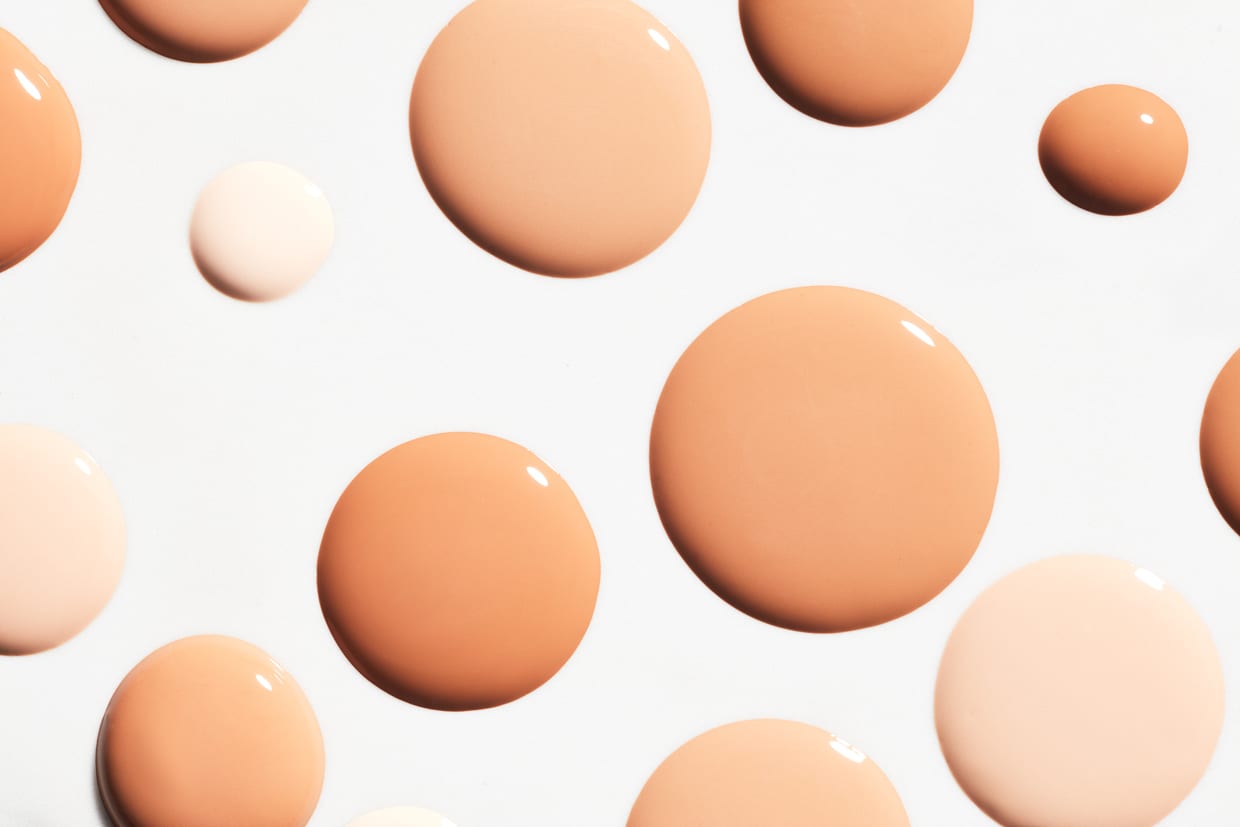 Overhead shot of drops of shades of beige liquid foundation on light gray background.