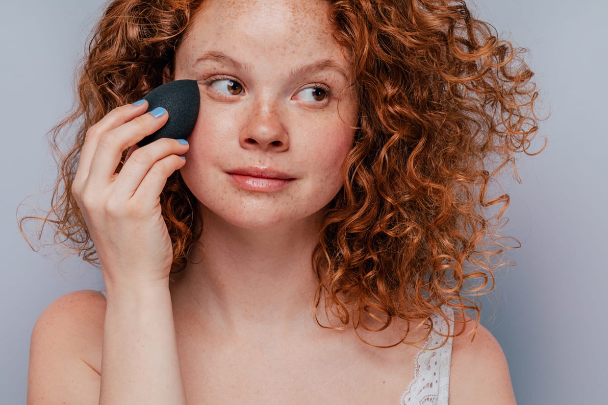 Close up of a woman applying foundation in front of a grey background.