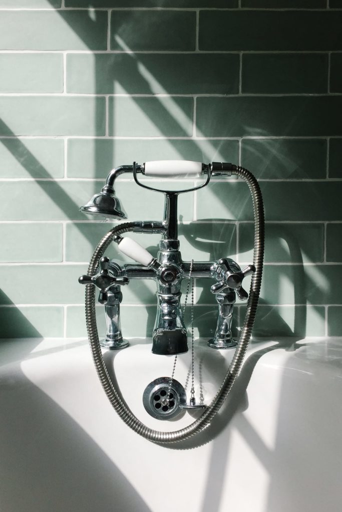 Bright sunlight shining through a window onto the faucet and shower head on a white enamel bath with light green tiles in the background.
