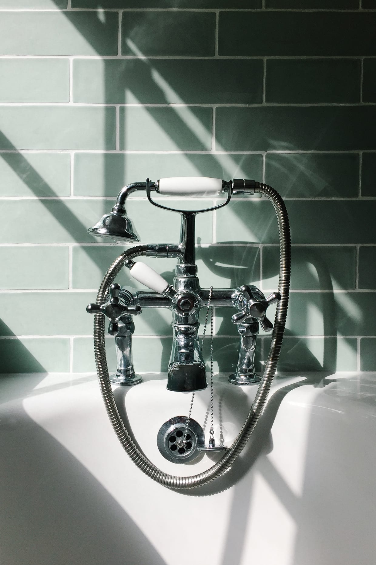 Bright sunlight shining through a window onto the faucet and shower head on a white enamel bath with light green tiles in the background.