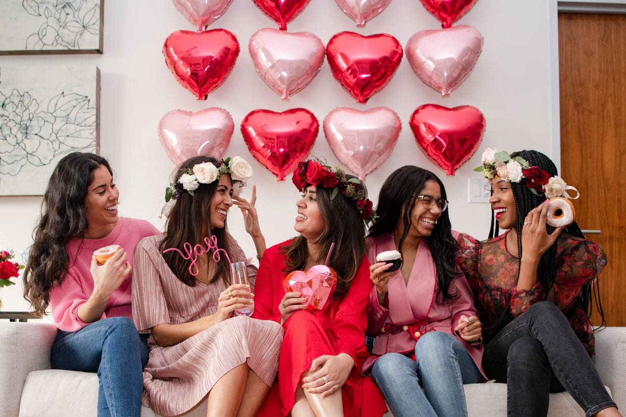 Four women dressed up in shades of red and pink sitting on a couch in front of a pink and red heart balloon wall.