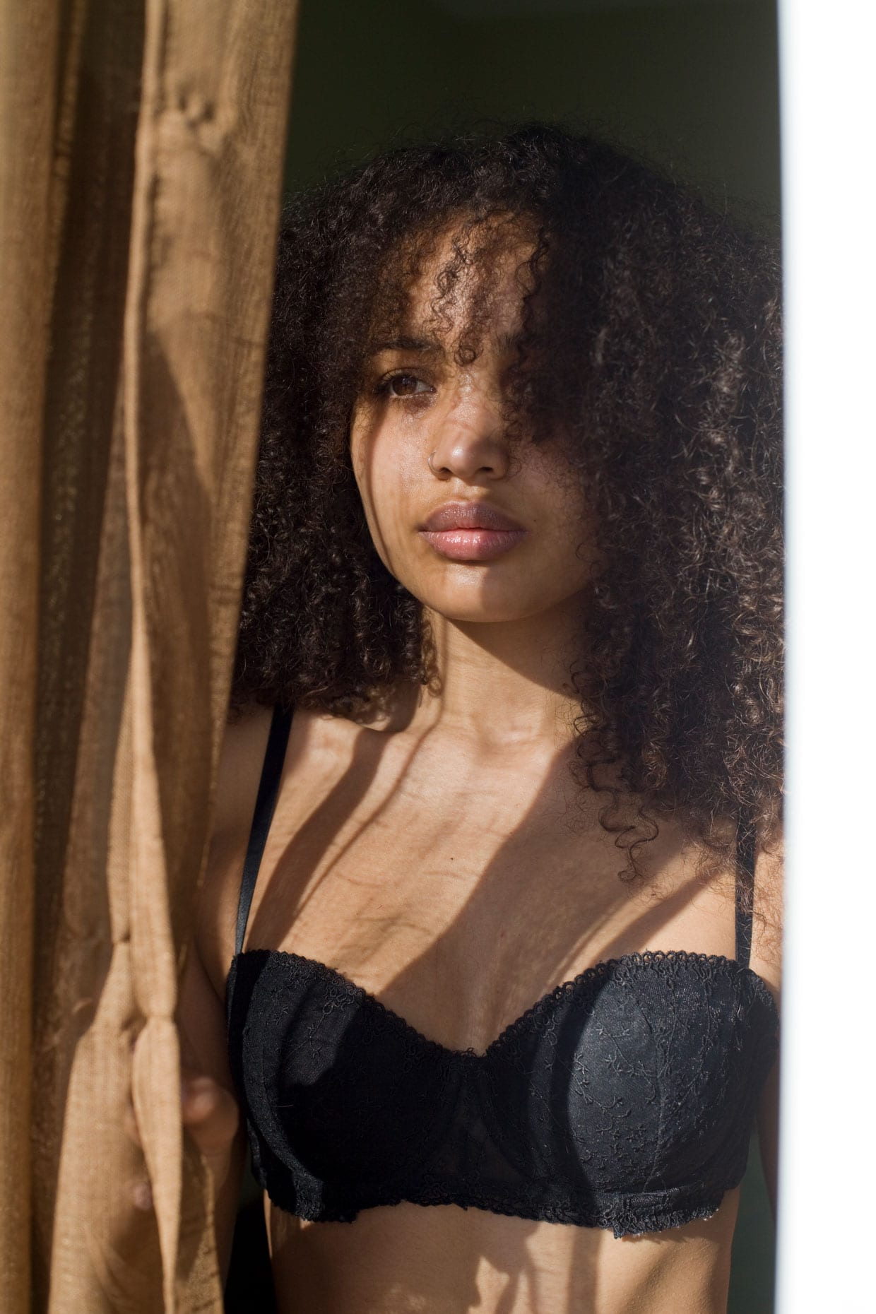 A young woman with curly brown hair, wearing a black bra, standing at a window, looking outside.