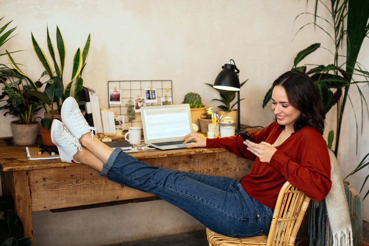 A woman using her phone in her home office that's full of plants.