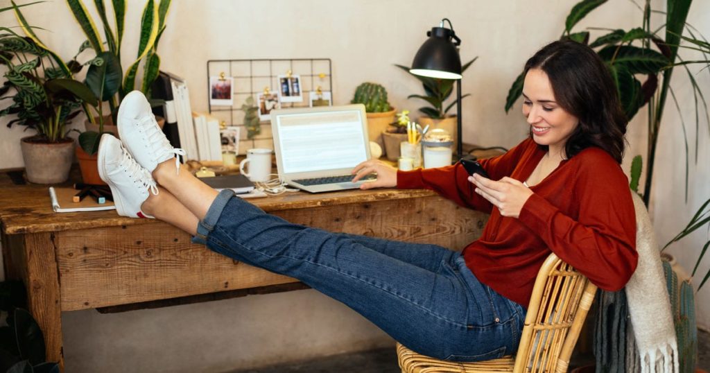 A woman using her phone in her home office that's full of plants.