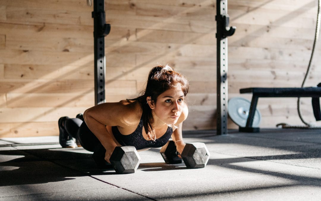 A woman holding dumbbells while doing push-ups with weights in a sunny, modern gym.