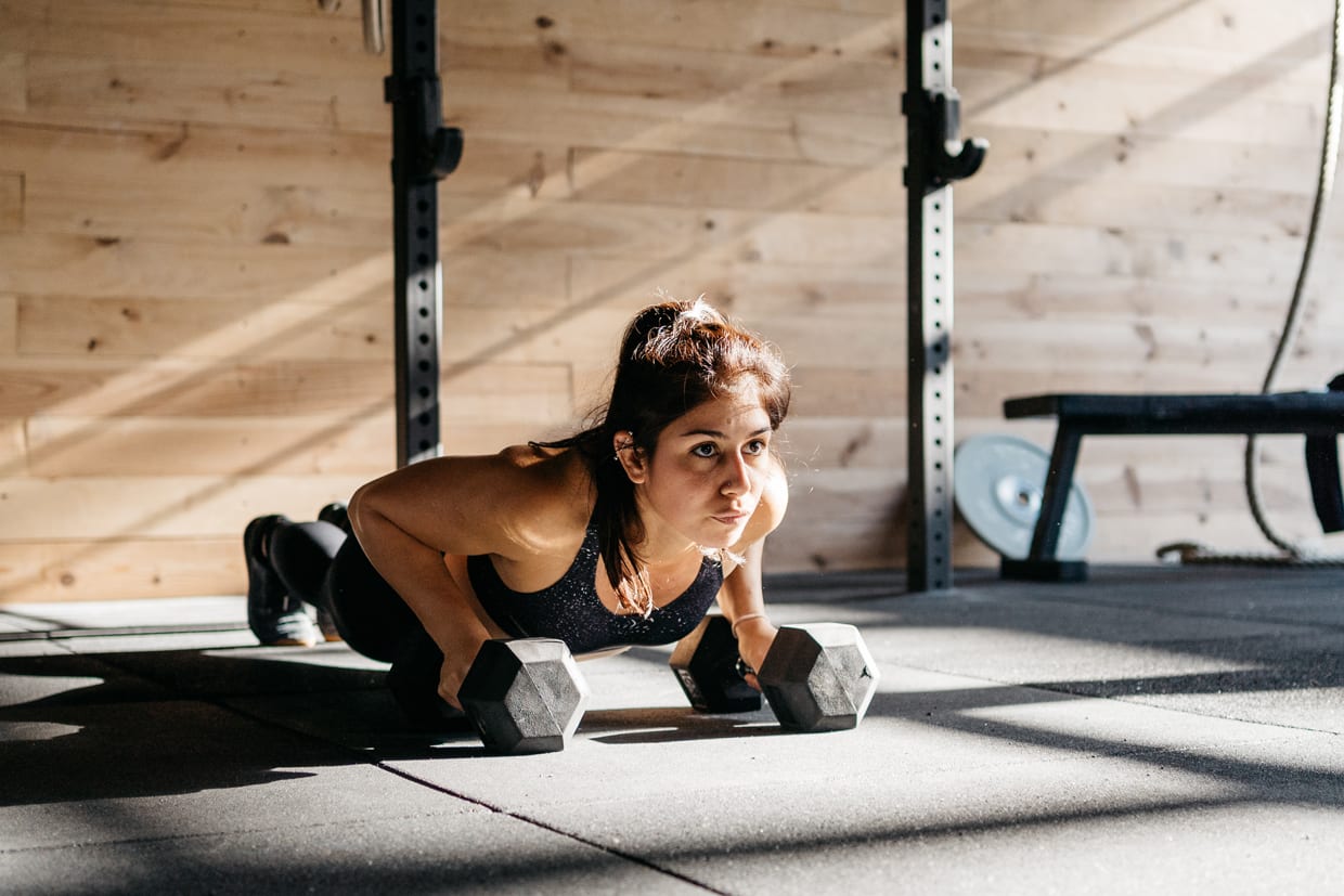 A woman holding dumbbells while doing push-ups with weights in a sunny, modern gym.