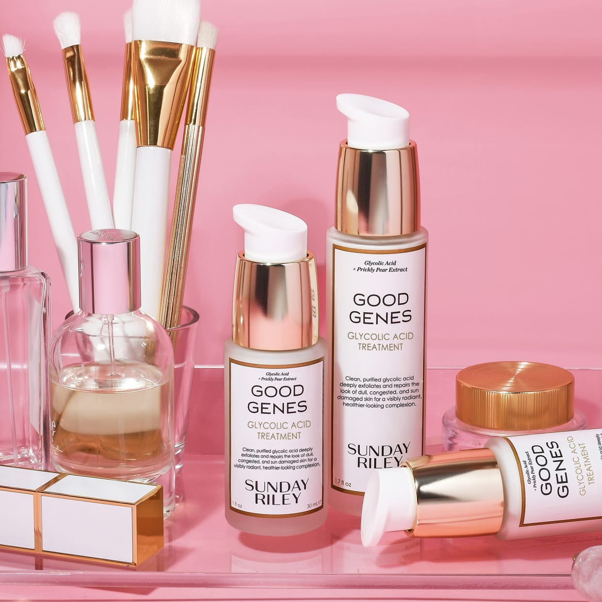 Multiple bottles of Sunday Riley Good Genes Glycolic next to an acrylic makeup brush holder filled with white and gold brushes on a pink background.
