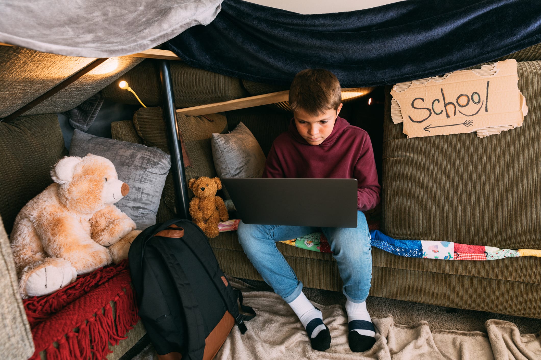 A young boy is working on his schooling and homework on a laptop from home. He sits inside a homemade couch fort trying his best to replicate his elementary education while at home during the coronavirus pandemic. Image taken in Utah, USA.