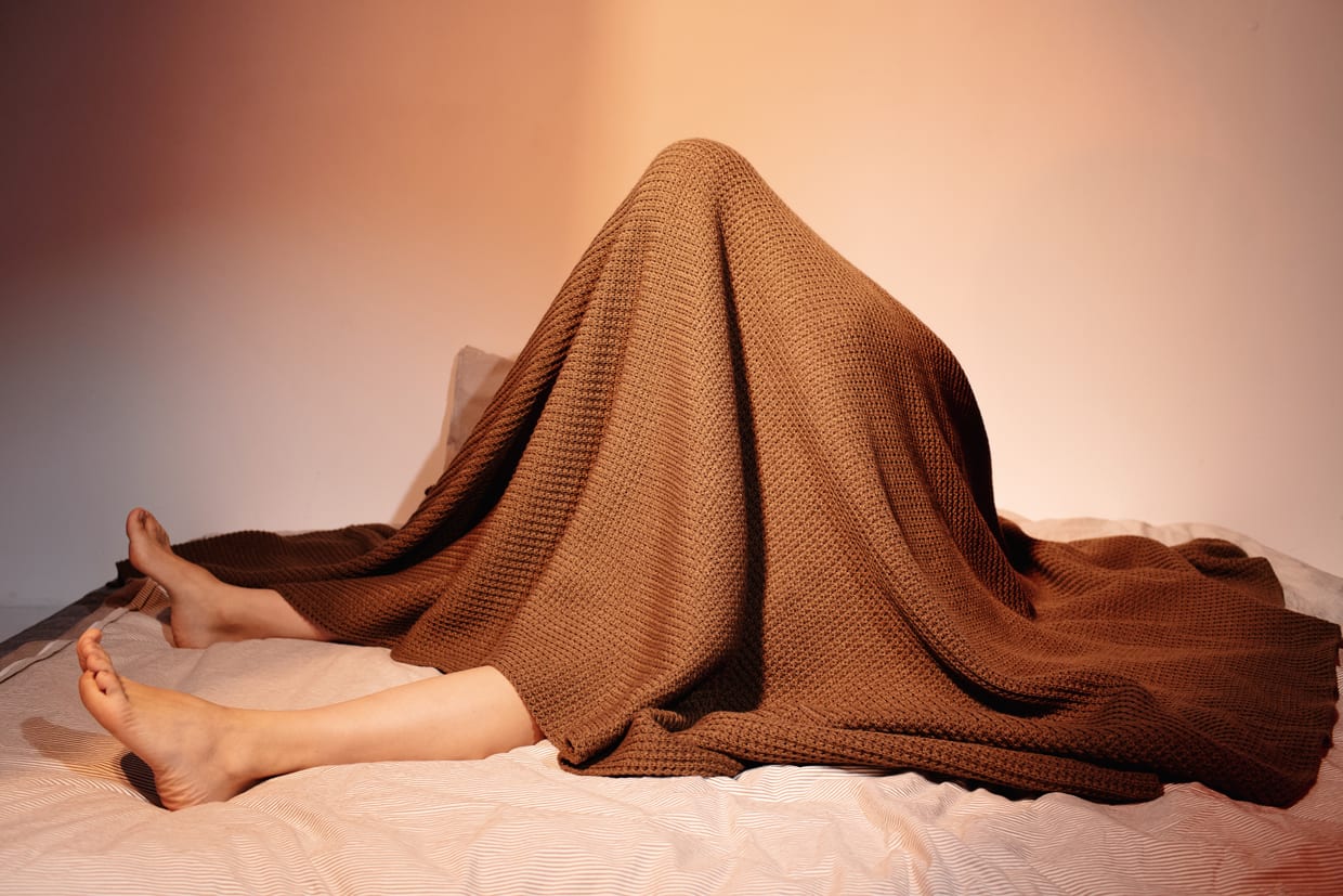 An unrecognizable woman hiding under brown blanket while sitting on a bed.