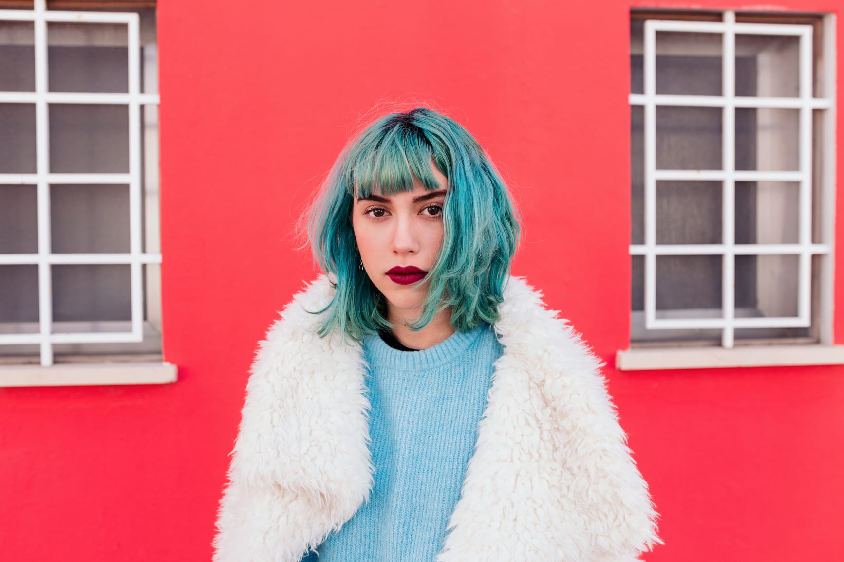 Cool young woman with blue hair wearing a white coat and looking at the camera with confidence. Red background behind.