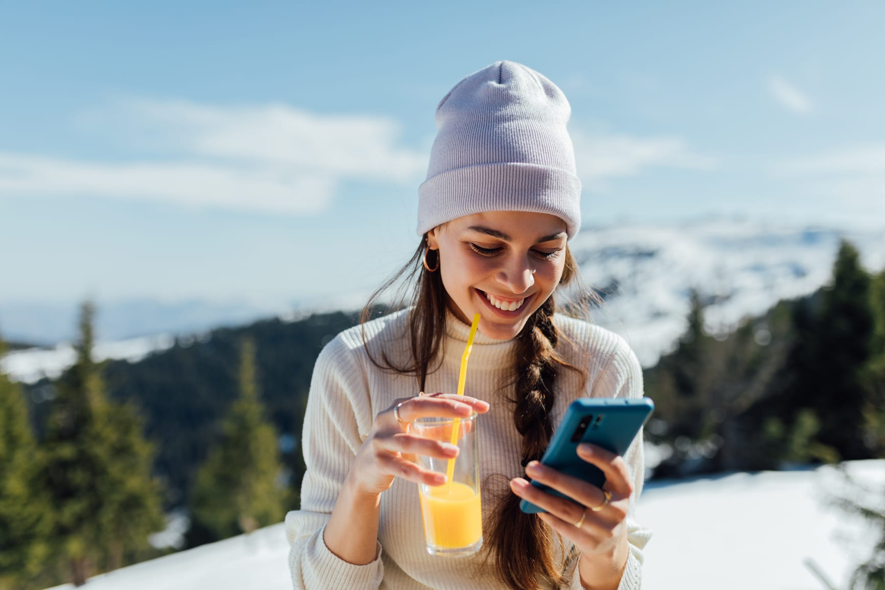 Young woman sitting outside in ski bar, using cellphone, drinking beverage and enjoying the sunny day.