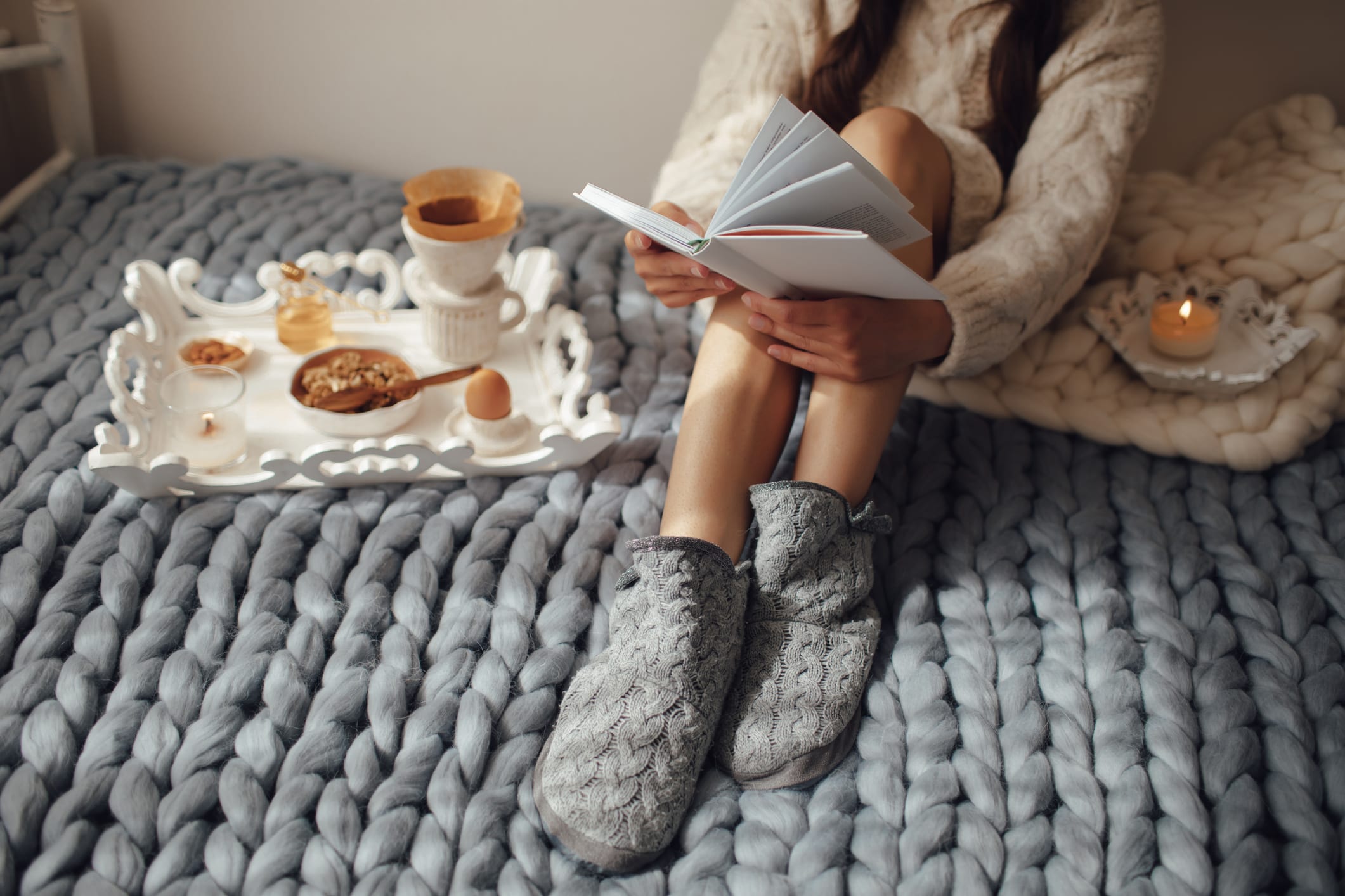 Woman with long hair drinking hot coffee and reading book in bed. Woman in woolen socks and sweater sitting on wool chunky merino plaid. Cozy winter morning concept.
